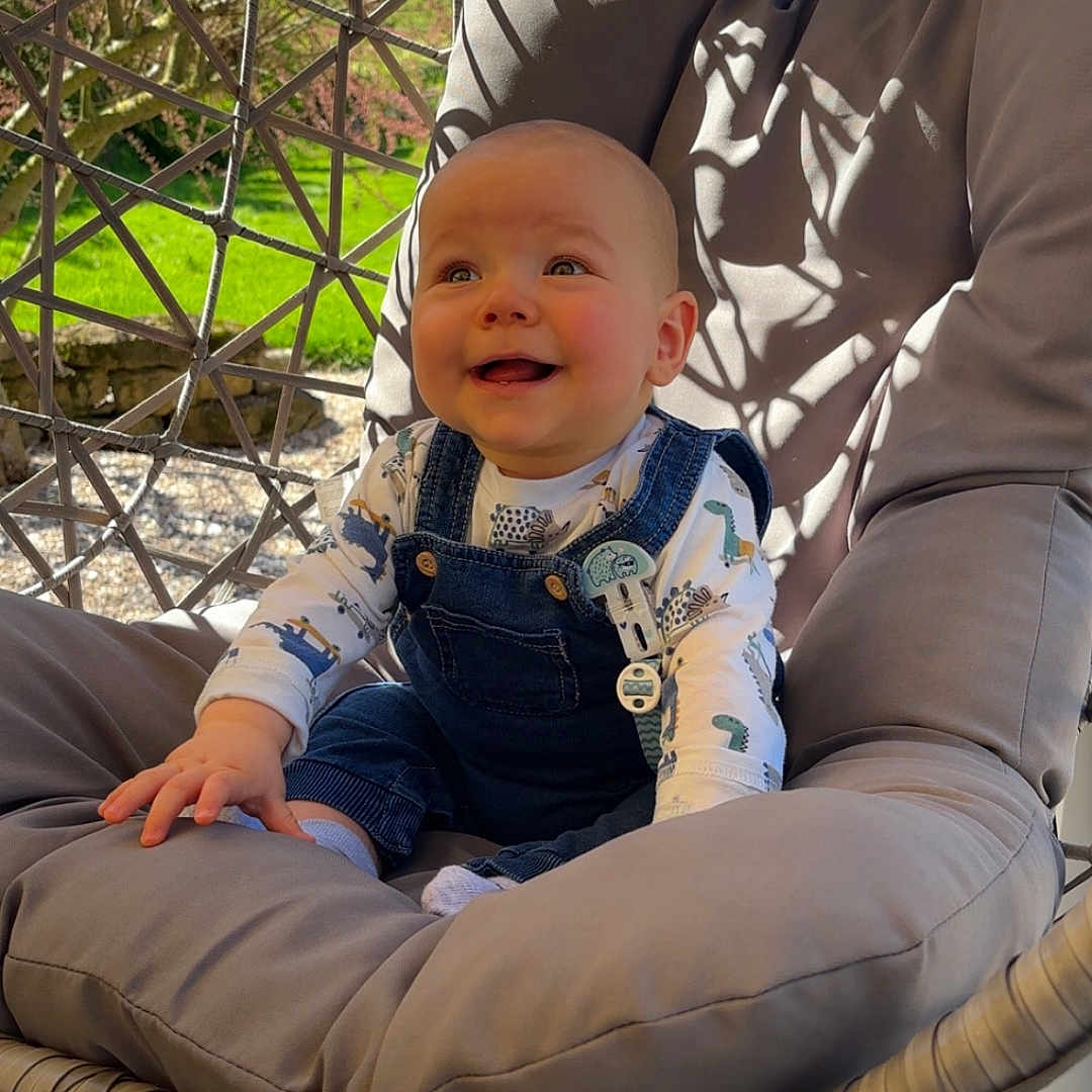 Sacha a rejoint le concours — aidez-le/la à gagner de superbes lots ! baby, casual_clothing, child, cushion, daylight, denim_overalls, dinosaur_print, garden, hanging_chair, happy, infant, outdoor, person, plush, portrait, relaxing, seat, smiling, sunlight, wooden_floor