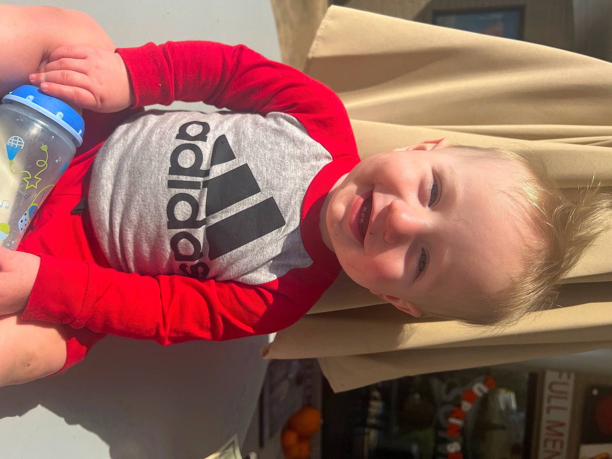 Ellis is registered to the contest to win money with this photo: baseball_cap, bottled_water, carmine, child, drinking_water, elbow, fun, happy, joy, knee, logo, mouth, nail, person, personal_protective_equipment, room, sleeve, smile, t_shirt, toddler