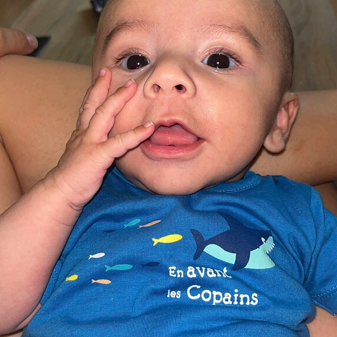 Eden participe au concours pour gagner de l'argent avec cette photo : baby, background, blue_shirt, child, clothing, cute, expression, eyes, face, fish_print, hand, indoors, infant, mouth, person, sitting, skin, table, text, tongue