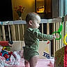 baby, child, onesie, playpen, toy, ring_toy, blanket, pink_blanket, indoor, wooden_bars, flower_vase, flowers, soft_toy, window, home, floor, standing, infant, toddler, cuddly