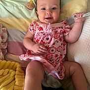 Taylynn is registered to the contest to win money with this photo: baby, child, infant, smile, dress, floral_pattern, blanket, cushion, pillow, indoors, happy, cute, person, lying_down, hand, face, head, soft_toys, colorful, cozy