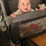 Nathaniel is registered to the contest to win money with this photo: baby, playpen, toys, smile, happy, indoor, child, face, hands, blanket, floor, wood_floor, furniture, mesh, clothing, person, infant, cute, expression, home