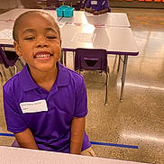 Michael is registered to the contest to win money with this photo: child, smiling, classroom, table, purple_polo, chair, school, indoor, person, education, happy, desk, floor, name_tag, writing, student, light, furniture, young, casual