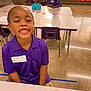 child, smiling, classroom, table, purple_polo, chair, school, indoor, person, education, happy, desk, floor, name_tag, writing, student, light, furniture, young, casual