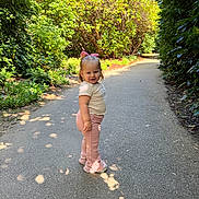 Athena is registered to the contest to win money with this photo: child, toddler, girl, standing, path, outdoor, sunlight, greenery, plants, shoes, bow, pink_pants, cream_top, nature, smile, portrait, daylight, casual_clothing, side_view, happy