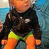 toddler, child, braids, beads, black_jacket, nike_logo, green_shorts, bed, blue_bedspread, indoor, person, sitting, young_child, curious_expression, casual_clothing, hands, legs, face, portrait, home
