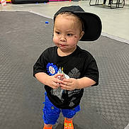 Austin is registered to the contest to win money with this photo: toddler, child, boy, cap, black_shirt, blue_shorts, orange_socks, juice_pouch, birthday_badge, indoor_play_area, play_structure, floor_mat, person, standing, expression, casual_clothing, indoor, woman, picnic_table, seated