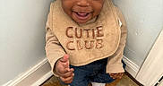 Joel is registered to the contest to win money with this photo: baby, child, smiling, happy, indoor, bib, clothing, curly_hair, carpet, corner, wall, sitting, person, cute, young_child, expression, portrait, footwear, home, toddler