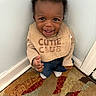 baby, child, smiling, happy, indoor, bib, clothing, curly_hair, carpet, corner, wall, sitting, person, cute, young_child, expression, portrait, footwear, home, toddler