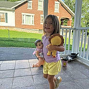 Kalani is registered to the contest to win money with this photo: brick_house, child, concrete_floor, daylight, doll, fence, girl, grass, happy, house, outdoor, playful, plush_toy, porch, purple_shirt, smiling, sneakers, toy, trees, yellow_shorts