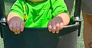 Elliott is registered to the contest to win money with this photo: adult_hand, baby, child, face, family, green_shirt, happy, infant, laughing, legs, outdoor, park, playground, portrait, safety_bar, shorts, sitting, smiling, stroller, sunlight