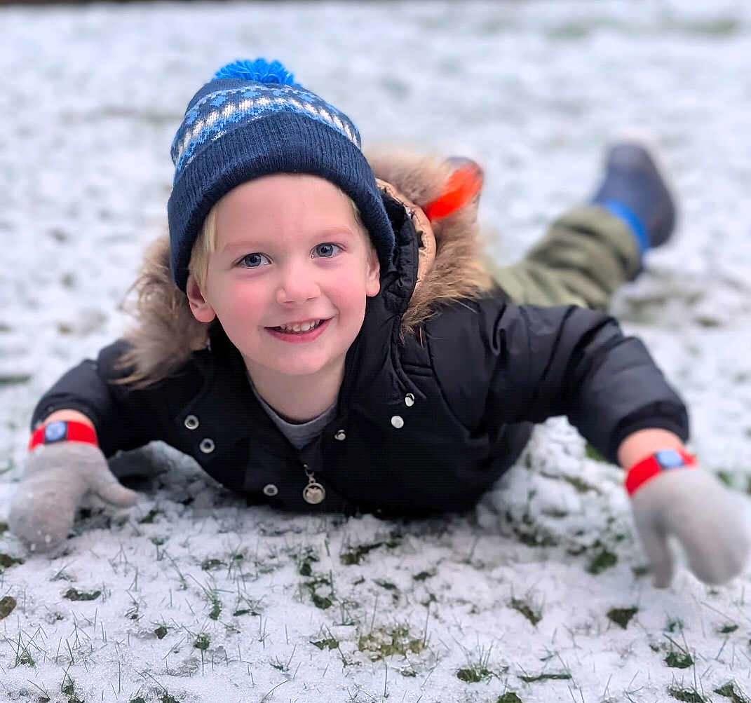 Owen is registered to the contest to win money with this photo: child, smiling, winter_hat, gloves, snow, grass, outdoor, cold_weather, jacket, happy, lying_down, blue_eyes, face, portrait, fun, playful, young, childhood, nature, seasonal
