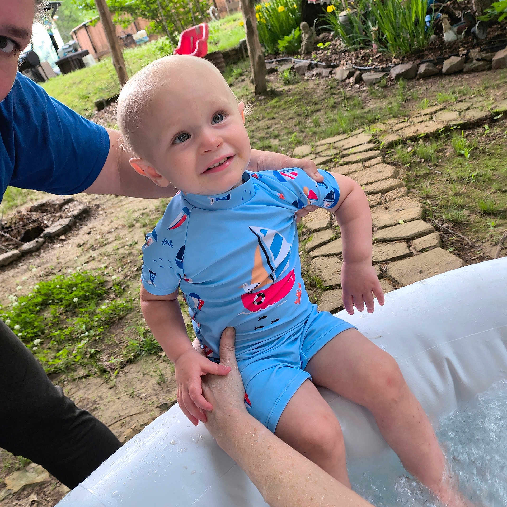 Remington is registered to the contest to win money with this photo: toddler, child, swimsuit, blue_clothing, inflatable_pool, adult_hand, outdoor, garden, grass, stone_path, water, summer, playtime, smiling, person, nature, sunlight, barefoot, happy, family