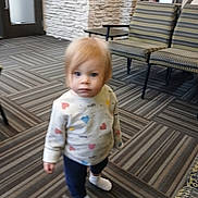 Clara joined the competition — help win amazing prizes! toddler, child, indoor, carpet, pattern, sweater, hearts, blonde_hair, white_socks, chair, furniture, stone_wall, flooring, person, young_child, curious, standing, casual_clothing, soft_lighting, blurry