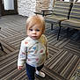 toddler, child, indoor, carpet, pattern, sweater, hearts, blonde_hair, white_socks, chair, furniture, stone_wall, flooring, person, young_child, curious, standing, casual_clothing, soft_lighting, blurry