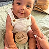 Tay is registered to the contest to win money with this photo: baby, infant, four_months, curly_hair, white_overalls, hand_holding, blanket, hat, cute, smiling, indoors, skin, foot, portrait, child, happy, sitting, person, close_up, family