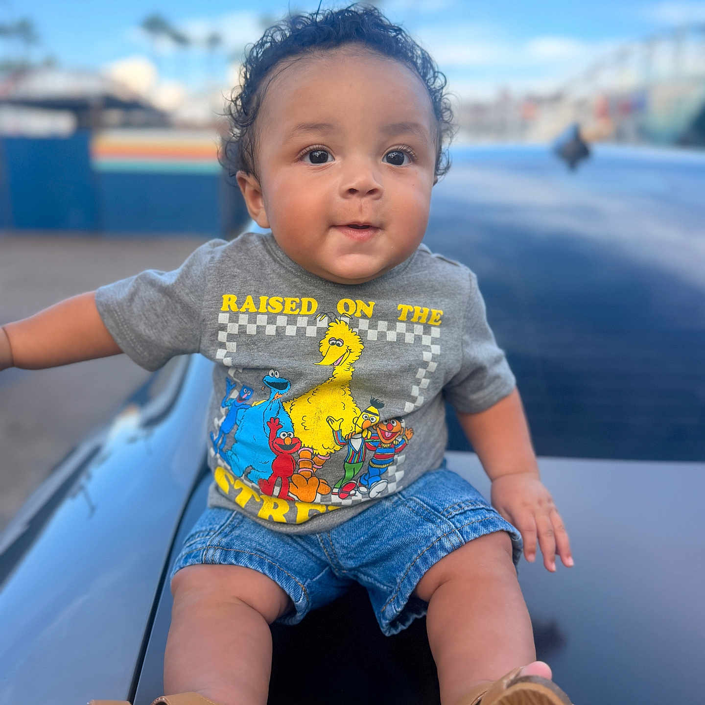 Tay joined the competition — help win amazing prizes! toddler, child, curly_hair, outdoor, blue_sky, denim_shorts, tshirt, sesame_street, footwear, sandals, face, portrait, sitting, smiling, casual_clothing, daylight, background_blur, person, cute, baby