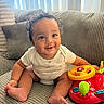 Tay is registered to the contest to win money with this photo: baby, child, curly_hair, smile, toy, colorful_toy, steering_wheel, couch, striped_couch, indoor, happy, sitting, onesie, infant, playful, furniture, hand, foot, window_blinds, bright