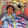 baby, child, curly_hair, smiling, playpen, colorful, pajamas, blue, yellow, toy, indoor, cute, happy, reaching_hand, patterned, fabric, floor, cushion, person, infant