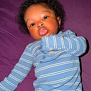 Kyrie is registered to the contest to win money with this photo: baby, blue_outfit, child, close_up, cozy, curly_hair, dark_skin, expression, face, flash_photography, indoor, infant, long_sleeves, looking_at_camera, lying_down, playful, portrait, purple_blanket, striped_clothing, tongue_out