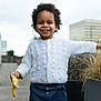 child, toddler, smiling, croissant, pastry, white_sweater, curly_hair, afro, standing, outdoor, rooftop, planter, ornamental_grass, urban, building, reflection, portrait, happy, food, hand