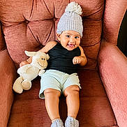 Skye is registered to the contest to win money with this photo: baby, child, hat, knitted_hat, booties, stuffed_animal, lamb, smiling, armchair, furniture, indoor, cozy, cute, happy, portrait, person, shorts, black_tank_top, plush_toy, relaxing