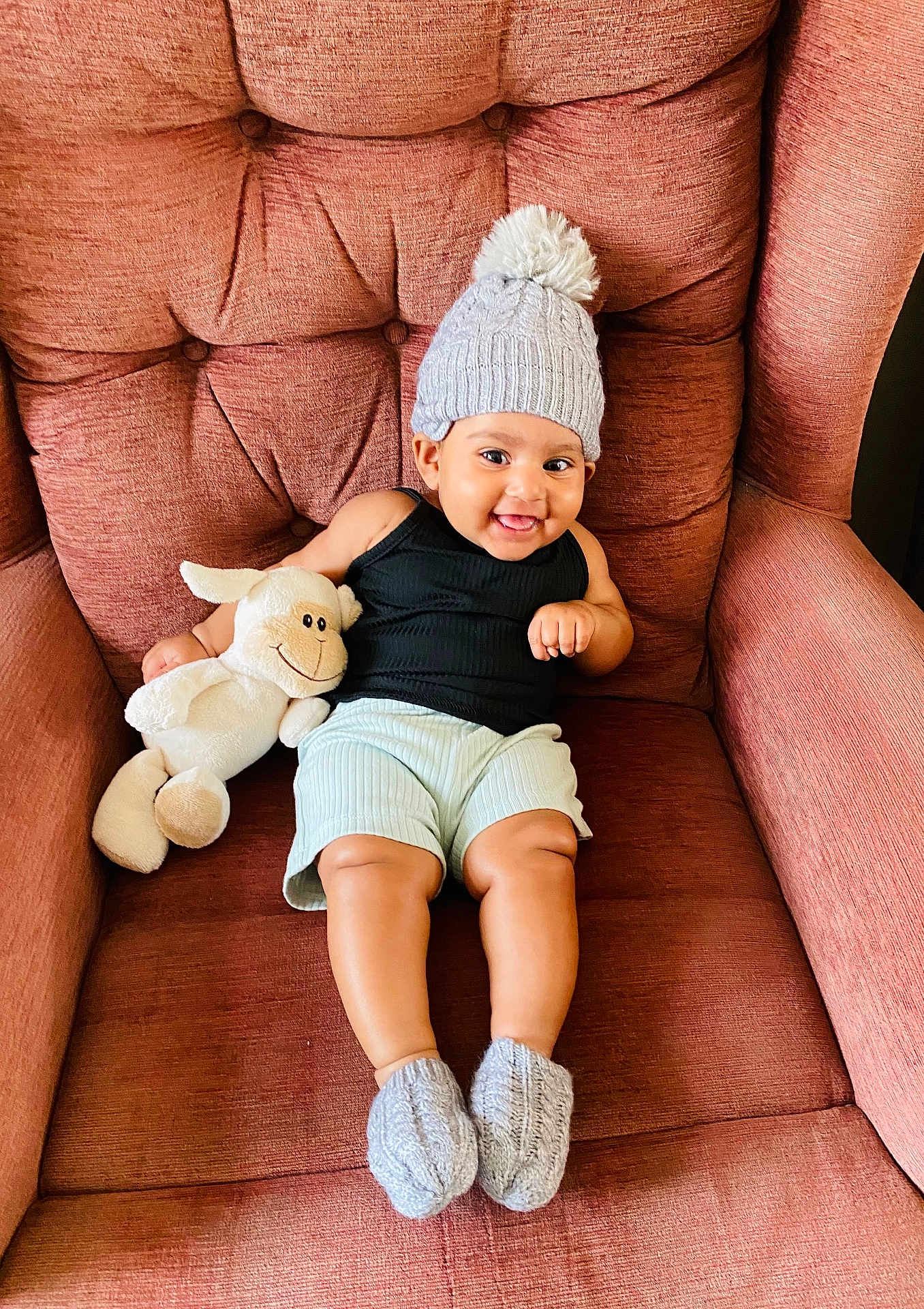 Skye is registered to the contest to win money with this photo: baby, child, hat, knitted_hat, booties, stuffed_animal, lamb, smiling, armchair, furniture, indoor, cozy, cute, happy, portrait, person, shorts, black_tank_top, plush_toy, relaxing