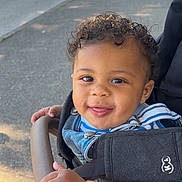 Kaïs participe au concours pour gagner de l'argent avec cette photo : toddler, child, curly_hair, smiling, stroller, outdoor, daylight, hands, clothing, denim, blue_stripes, happy, face, baby, person, seatbelt, pavement, sunlight, grip, cute