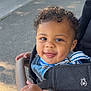 toddler, child, curly_hair, smiling, stroller, outdoor, daylight, hands, clothing, denim, blue_stripes, happy, face, baby, person, seatbelt, pavement, sunlight, grip, cute