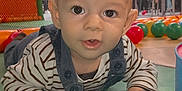 éden participe au concours pour gagner de l'argent avec cette photo : baby, child, indoor, playmat, colorful_balls, mesh_barrier, striped_shirt, overalls, curious_expression, hands, floor, people_in_background, red_chair, green, yellow_ball, smiling, toddler, play_area, person, head