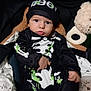 baby, child, costume, hat, plush_toy, cushion, blanket, seat, indoors, portrait, cute, black_clothing, white_clothing, green_splatter, infant, face, person, hands, expression, cozy