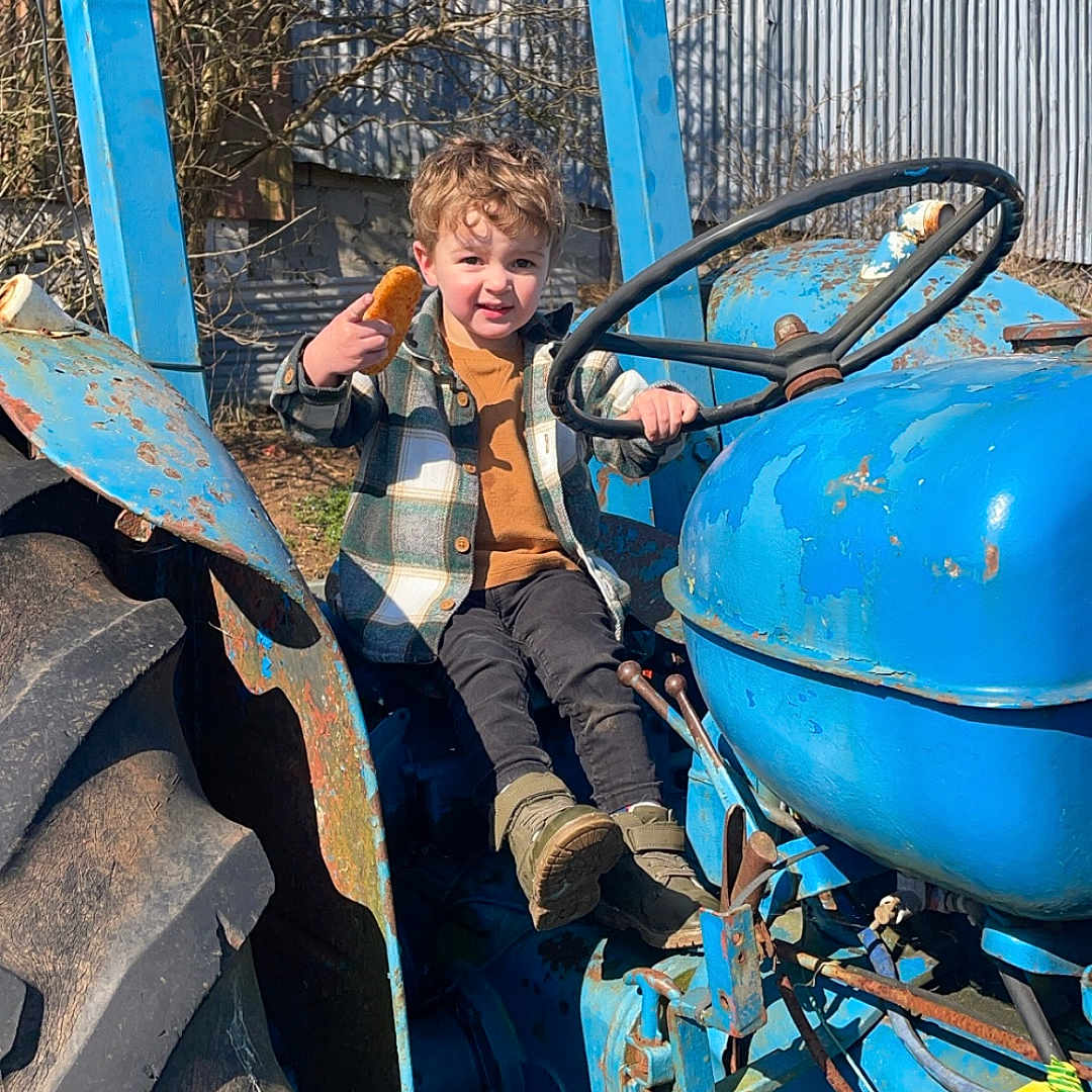 Greyson is registered to the contest to win money with this photo: blue, boots, child, clothing, daytime, fence, jacket, metal, outdoor, person, playful, rust, rusty, snack, steering_wheel, sunny, tire, tractor, tree, vehicle