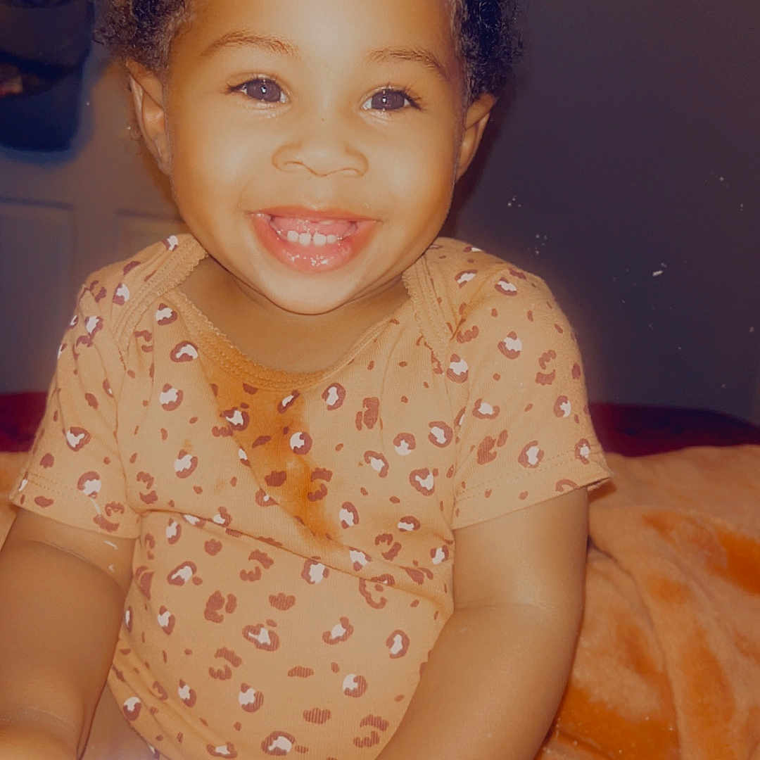 Journei is registered to the contest to win money with this photo: baby, baby_bottle, child, cozy, curly_hair, cute, face, happy, indoors, infant, leopard_print_clothing, orange_blanket, portrait, sitting, skin, smiling, toddler, two_buns, warm_lighting, young_child