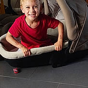 Gabriel participe au concours pour gagner de l'argent avec cette photo : child, boy, smiling, red_shirt, car_seat, toy_car, indoor, floor, happy, grip, blond_hair, casual_clothing, playful, seat, person, young, cute, childhood, portrait, fun