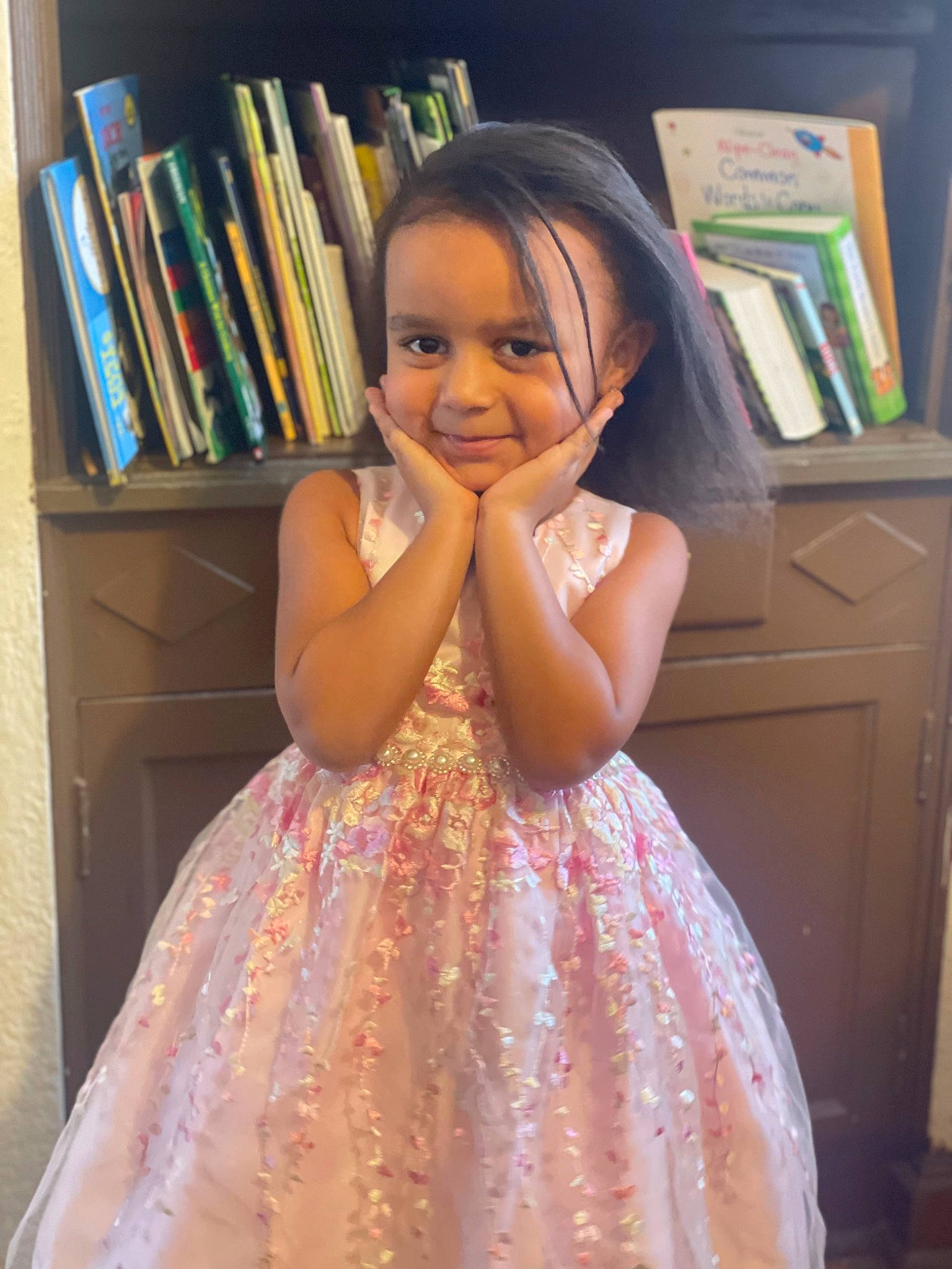 Azari is registered to the contest to win money with this photo: book, bookcase, child, day_dress, dress, fashion_design, fun, gown, hair, happy, human_body, iris, joy, one_piece_garment, person, pink, shelving, smile, standing, toddler
