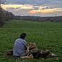 casual_clothing, clouds, companion, dog, evening, field, grass, hills, landscape, leisure, man, nature, outdoor, pet, relaxation, scenery, sitting, sky, sunset, trees
