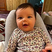 Charlotte joined the competition — help win amazing prizes! baby, child, smiling, face, person, indoor, clothing, floral_pattern, onesie, hand, lap, wood_paneling, furniture, cushion, happy, portrait, infant, skin, hair, cozy