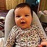 baby, child, smiling, face, person, indoor, clothing, floral_pattern, onesie, hand, lap, wood_paneling, furniture, cushion, happy, portrait, infant, skin, hair, cozy