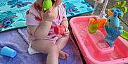 Sara is registered to the contest to win money with this photo: baby, child, towel, toy, water_playset, outdoor, grass, blanket, sunlight, concrete, garden, legs, blue_eyes, pink_clothing, green_toy, red_toy, playtime, happy, summer, person
