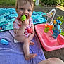 baby, child, towel, toy, water_playset, outdoor, grass, blanket, sunlight, concrete, garden, legs, blue_eyes, pink_clothing, green_toy, red_toy, playtime, happy, summer, person