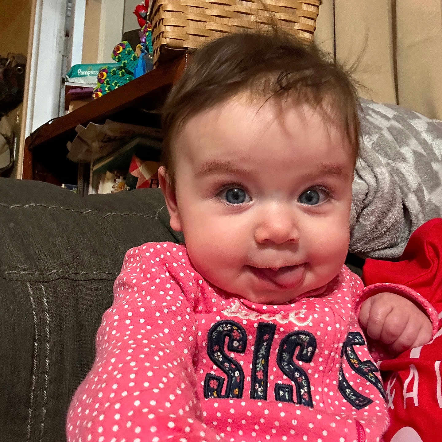 Arya is registered to the contest to win money with this photo: baby, child, face, tongue_out, blue_eyes, pink_clothing, polka_dot, cute, indoor, living_room, basket, expression, person, portrait, smiling, sitting, hair, hand, cozy, home