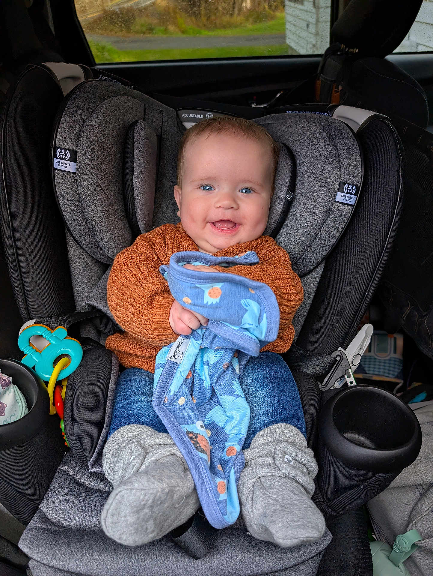 Samson joined the competition — help win amazing prizes! baby, car_seat, child, blue_eyes, orange_sweater, blanket, gray_booties, smiling, indoor, seatbelt, toy, car_interior, cute, infant, happy, clothing, portrait, holding, cozy, comfort
