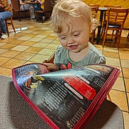 Kinleigh is registered to the contest to win money with this photo: blonde_hair, brown_chair, casual_clothing, child, curious, curly_hair, dining, family, floor_tiles, food_menu, indoor, menu, person, reading, restaurant, seated, smiling, table, toddler, wooden_chairs