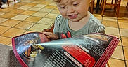 Kinleigh is registered to the contest to win money with this photo: blonde_hair, brown_chair, casual_clothing, child, curious, curly_hair, dining, family, floor_tiles, food_menu, indoor, menu, person, reading, restaurant, seated, smiling, table, toddler, wooden_chairs