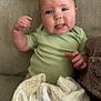 baby, child, tongue_out, green_onesie, blanket, couch, soft_texture, face, hand, infant, cute, indoor, portrait, skin, expression, sitting, young_child, comfort, cozy, furniture