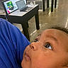 Kyren is registered to the contest to win money with this photo: baby, blue_clothing, casual, child, close_up, curious, expression, eyes, face, holding, human, indoor, infant, laptop, looking_up, person, portrait, skin, table, technology_store