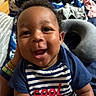 Kyren is registered to the contest to win money with this photo: baby, bed, blue_shirt, child, clothing, curly_hair, cute, fabric, face, hand, happy, indoor, infant, person, pillow, portrait, relaxed, smiling, striped_shirt, text_on_clothing