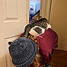 Fuzzy is registered to the contest to win money with this photo: animal, beanie, bent_over, casual, cat, cozy, door, feline, indoor, lighting, long_hair, maroon_shirt, pajama_pants, patterned_pants, person, pet, room, tabby_cat, white_cat, wood_floor
