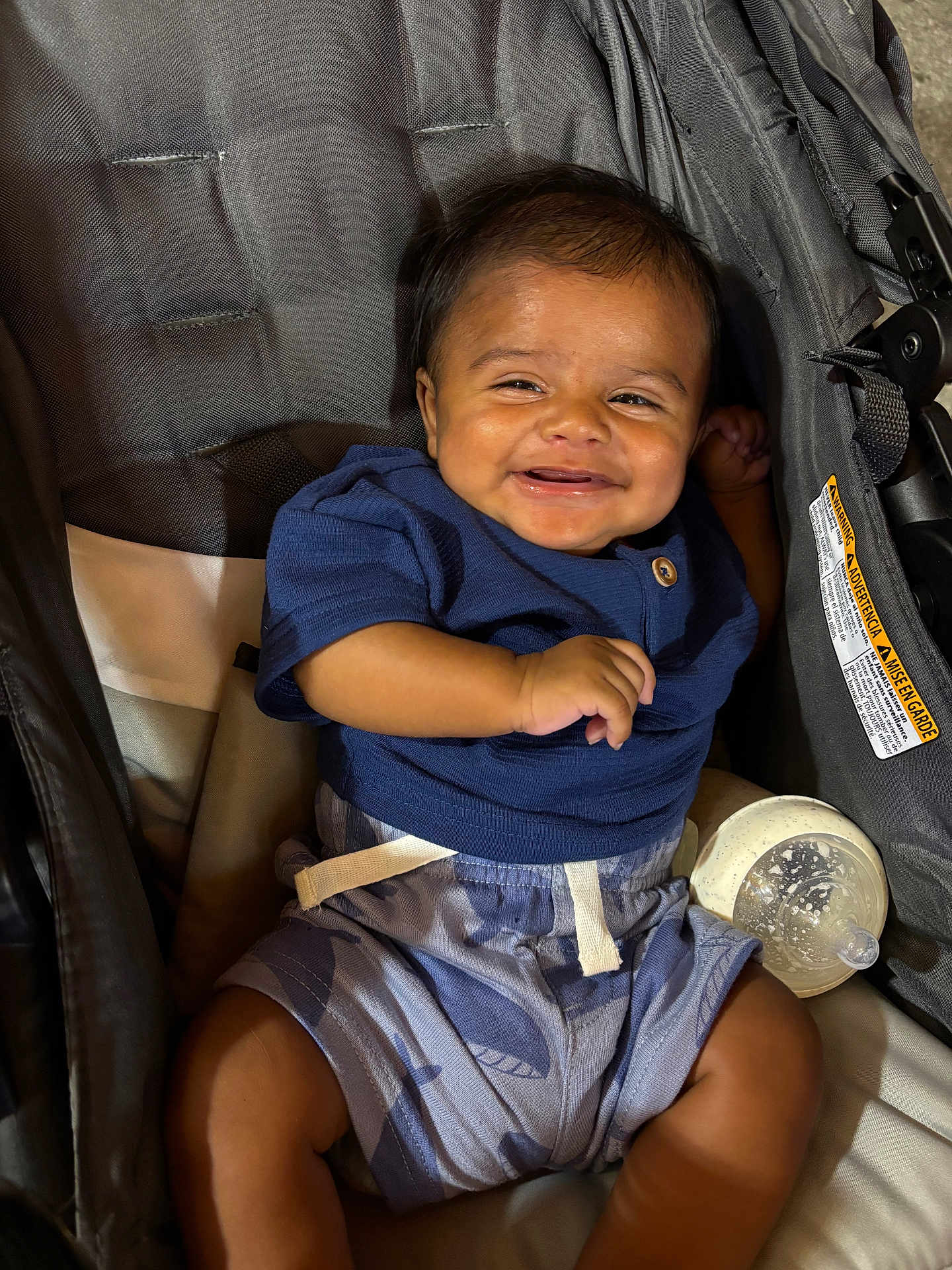 Frankie is registered to the contest to win money with this photo: baby, child, smiling, stroller, bottle, blue_shirt, shorts, infant, happy, portrait, person, seatbelt, fabric, comfort, cute, young, sitting, indoors, cheerful, casual