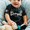 baby, smiling, black_headband, plush_toy, pacifier, blanket, child, indoor, portrait, happy, sitting, jersey, mexican_flag, cute, infant, soft_texture, cozy, headwear, toy, candid